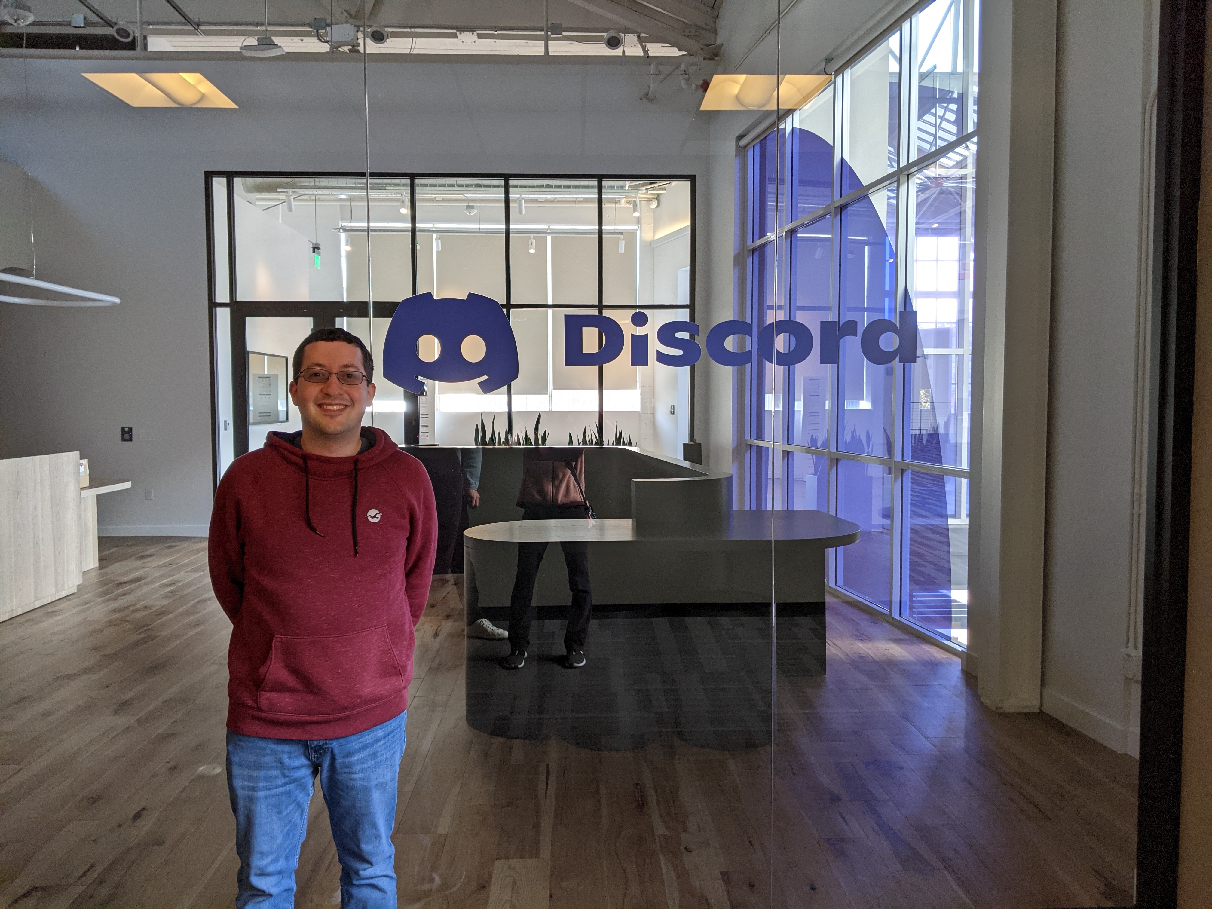 Ross, grinning, standing in front of a glass wall with the Discord logo. Behind the glass is an empty reception desk.